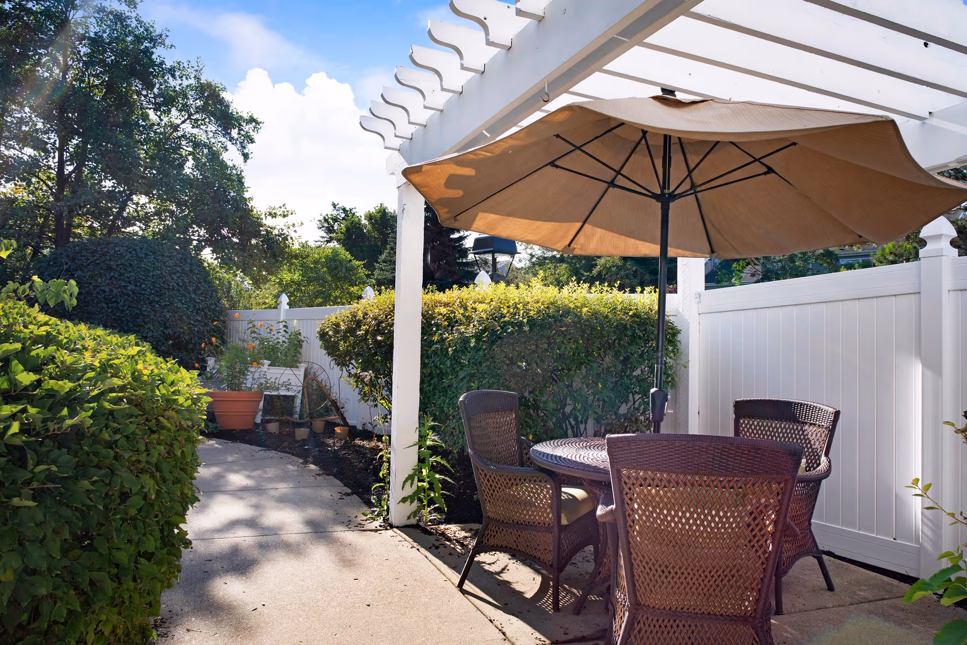 Outdoor patio with a round wicker table, four chairs and a large umbrella under a white pergola next to shrubs and a white fence.