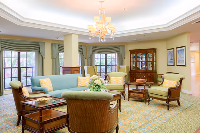 A bright and spacious living room area with a light blue sofa, several green armchairs with yellow patterned cushions, wooden side tables, a floral centerpiece on a coffee table, large windows with green curtains, a chandelier hanging from a tray ceiling, and a wooden china cabinet against the wall.