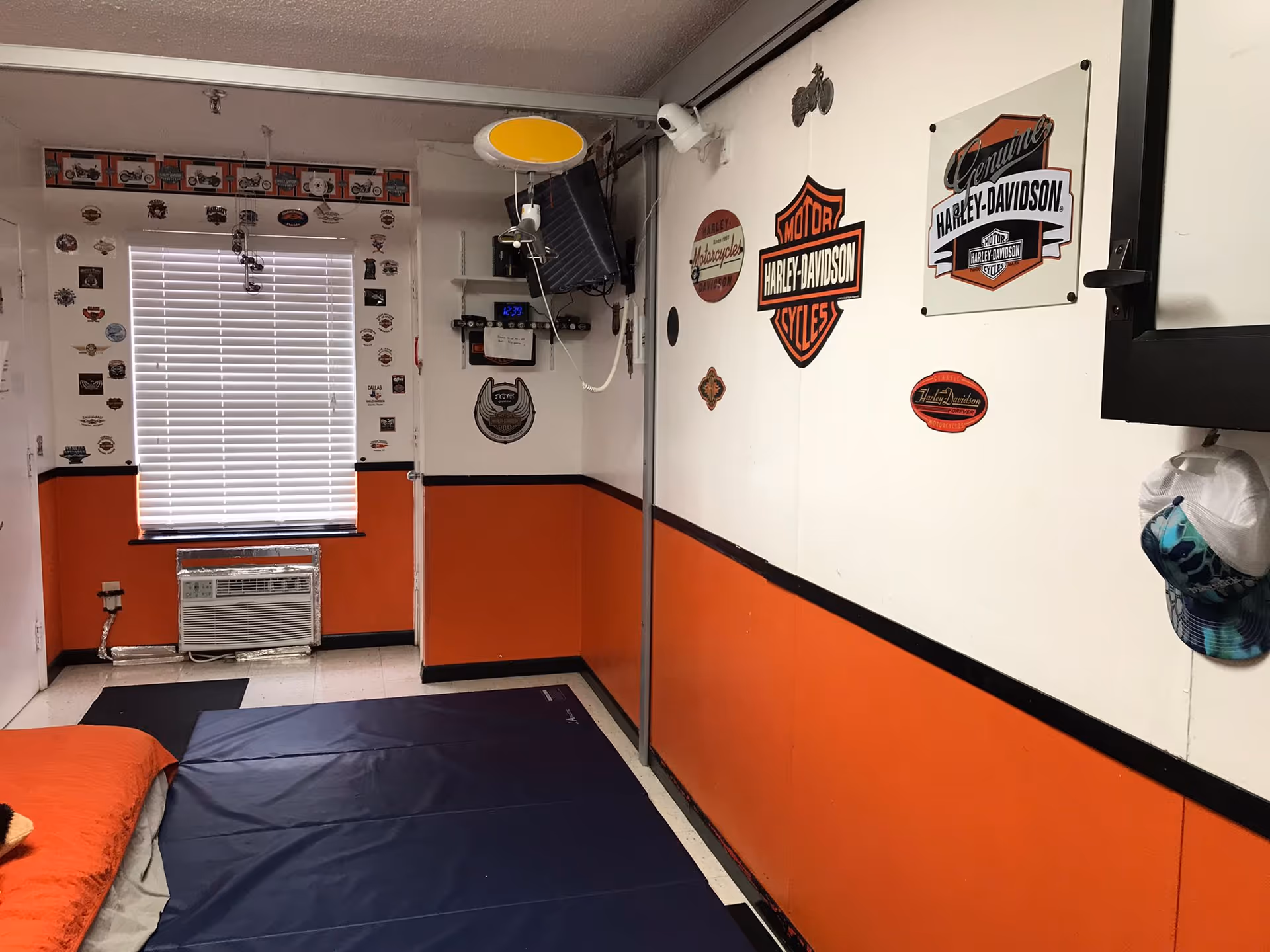 Small interior room with orange lower walls decorated with Harley-Davidson signs, a window with blinds, a wall-mounted TV, and a mattress/mat on the floor.