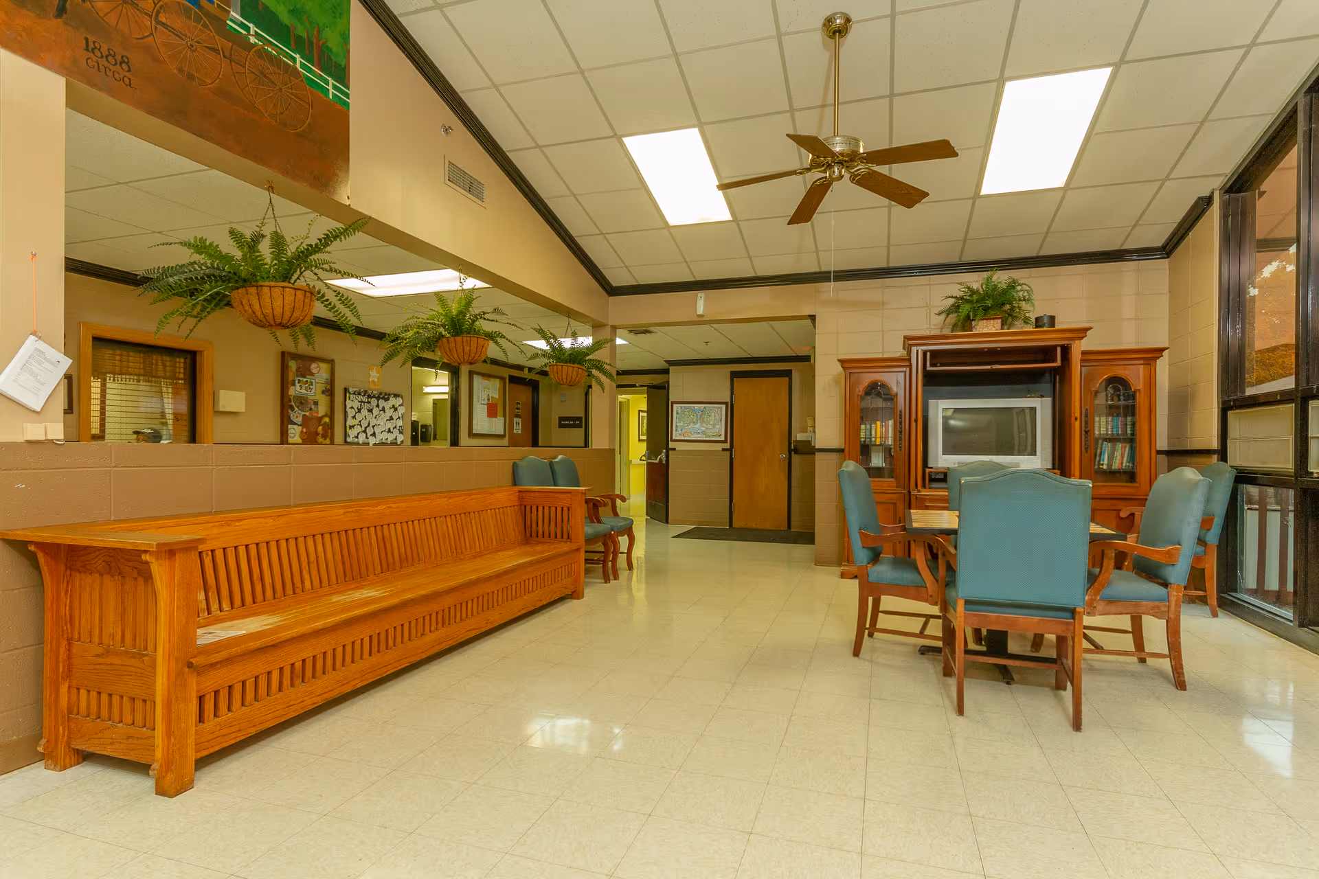 Bright common room with a long wooden bench, a table and upholstered chairs, a TV cabinet, hanging plants and a ceiling fan.