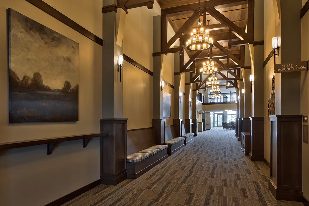 A spacious hallway with high ceilings featuring wooden beams and multiple chandeliers. The walls are adorned with paintings and wall sconces. There are built-in wooden benches with cushions along the sides. A sign indicates the direction to the Chapel / Community Room. The far end of the hallway has large windows letting in natural light.