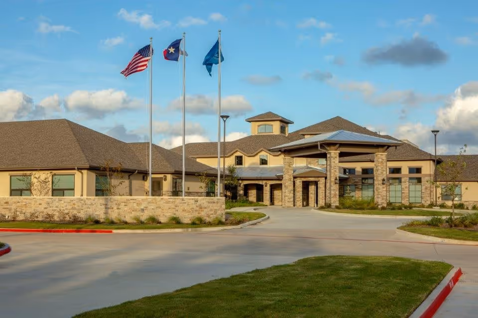 Exterior view of The Shores at Clear Lake facility showing a large building with a stone facade and multiple peaked roofs under a partly cloudy sky. Three flagpoles with the American flag, Texas state flag, and another flag stand in front of the building. There is a driveway and landscaped grass areas in the foreground.