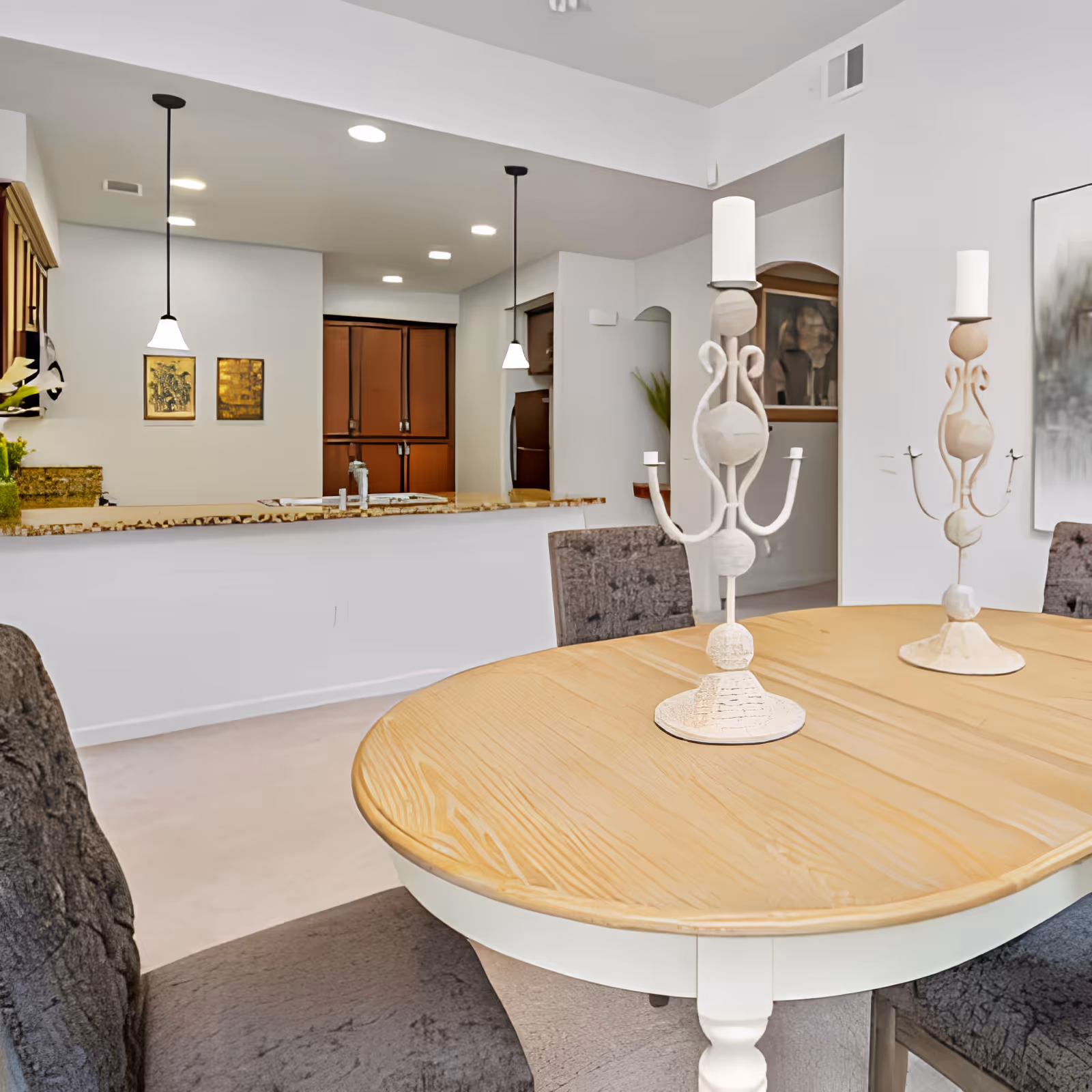 A dining area with a wooden oval table and four upholstered chairs. Two decorative white candle holders are placed on the table. In the background, there is a kitchen with a granite countertop, pendant lights hanging from the ceiling, and wooden cabinets. The walls are white, and there are framed pictures on the wall near the kitchen.