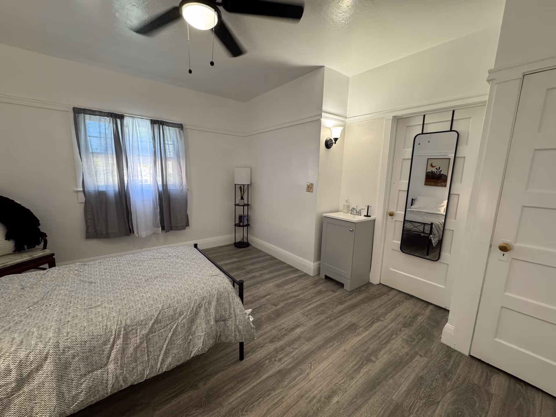 Simple bedroom with a bed, ceiling fan, window with curtains, a floor lamp, a small sink cabinet, and a full-length mirror hanging on a white door.