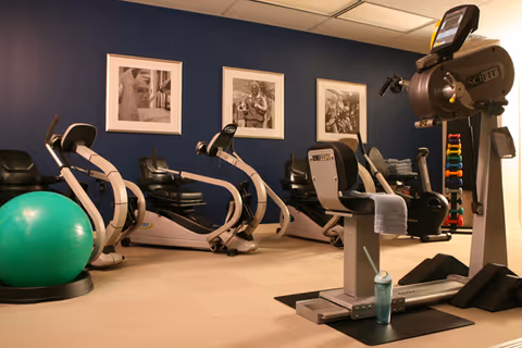 Indoor exercise room with several recumbent stationary bikes, a large green exercise ball, a set of colorful dumbbells on a rack, and a water bottle on the floor. The walls are painted dark blue and decorated with three framed black and white photographs.