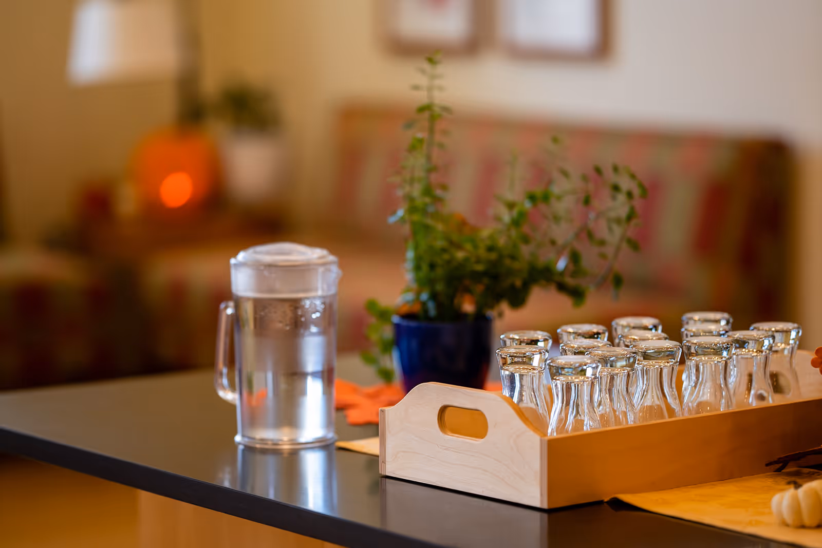 A countertop with a water pitcher, a wooden tray of glasses, and a potted plant in a blurred common room.