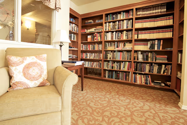 Cozy reading area with an upholstered armchair, side table and lamp in front of wall-to-wall bookshelves in a carpeted room.