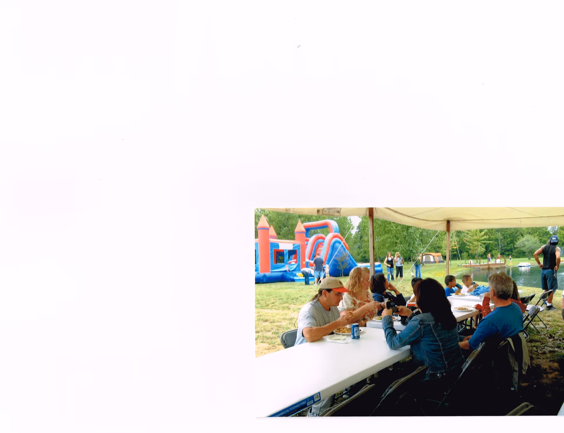 A group of people sitting at a long white folding table under a canopy tent outdoors, eating and socializing. In the background, there is a colorful inflatable bounce house and slide, green trees, and a small body of water with people near the shore.