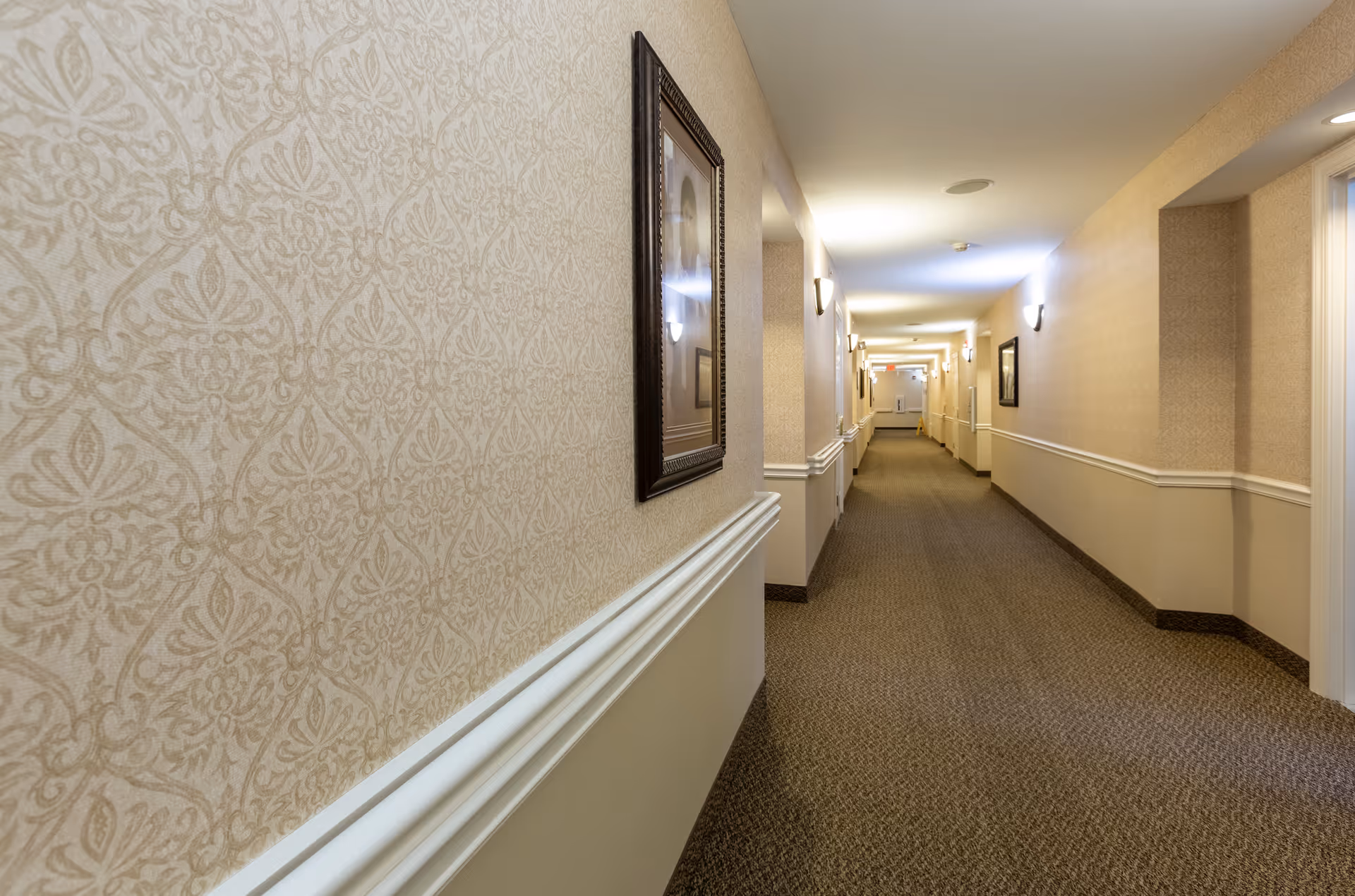 A long, well-lit hallway in a senior living facility with patterned wallpaper, carpeted floor, framed pictures on the walls, and wall-mounted light fixtures.
