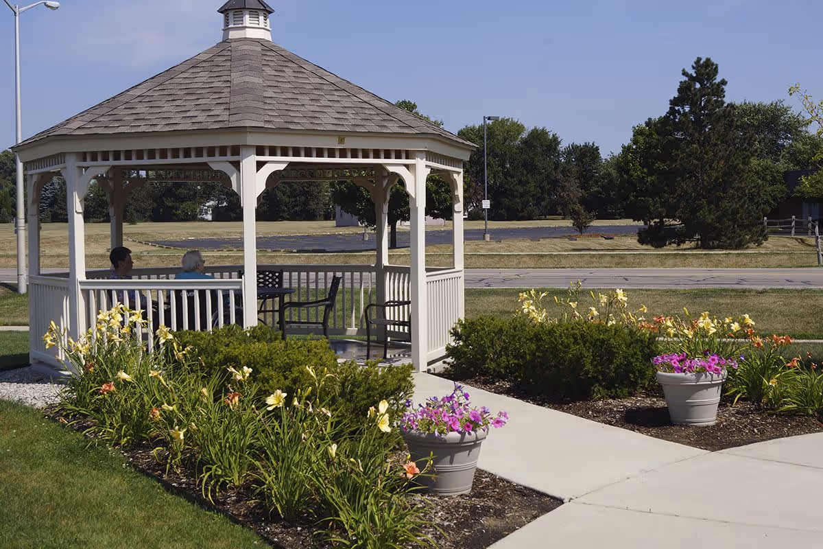 A white gazebo with a shingled roof in a garden area with blooming flowers and shrubs. Two people are sitting inside the gazebo on black chairs. The background shows a road, trees, and a clear blue sky.