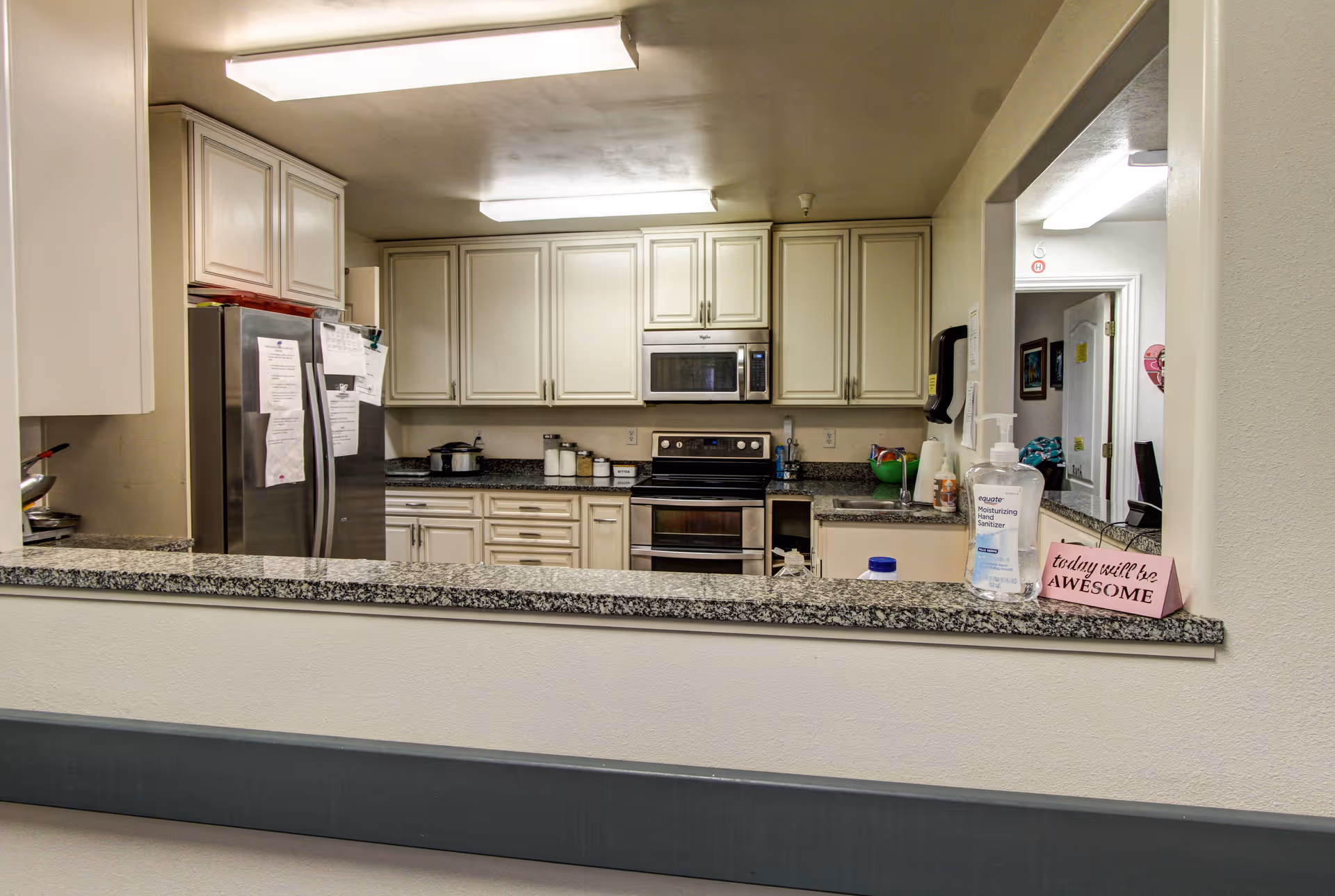 Open kitchen with granite pass-through counter, white cabinets, stainless steel refrigerator and stove, and hand sanitizer on the counter.