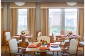 A bright dining room with several round wooden tables, each set with orange napkins and floral centerpieces. The room has large windows with beige curtains allowing natural light to fill the space. Comfortable upholstered chairs surround the tables.