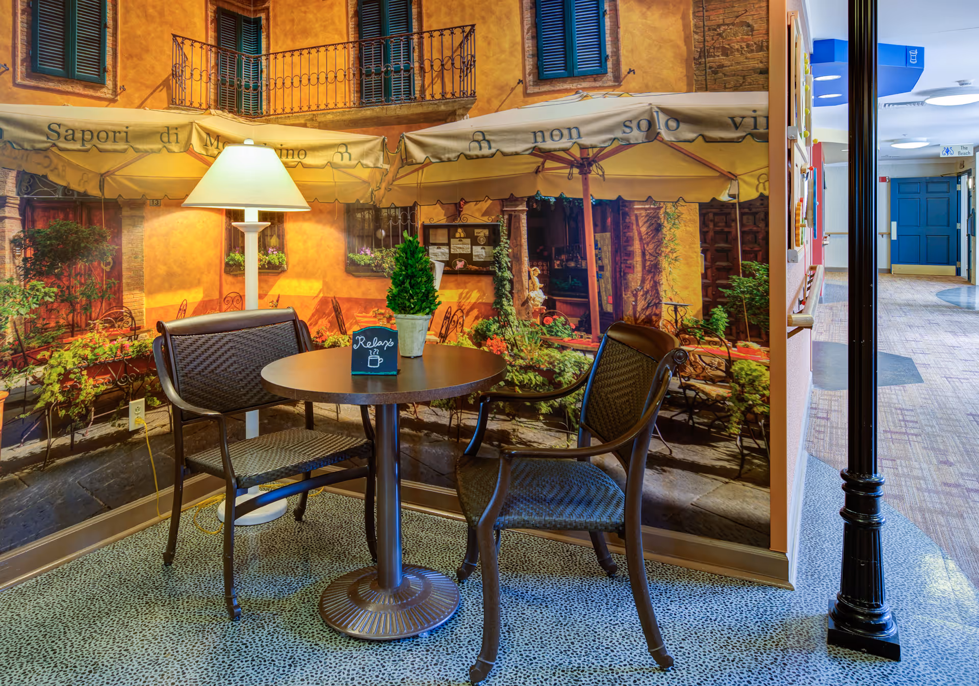 A small seating area with a round table and two chairs in front of a wall mural depicting an outdoor café scene with yellow walls, green shutters, and umbrellas. A floor lamp stands next to the table, which has a small sign that says 'Relax' and a potted plant on it. The floor has a speckled pattern, and a hallway with blue doors is visible to the right.