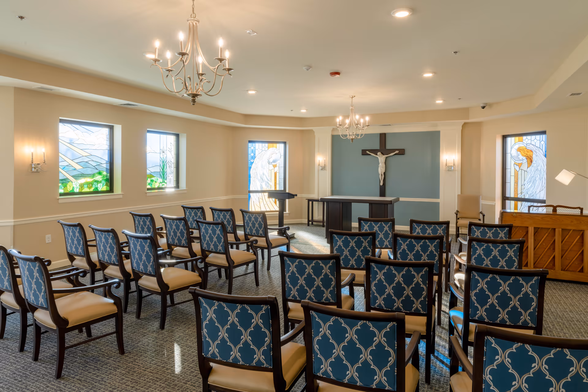 A small chapel room with rows of blue patterned chairs facing an altar with a crucifix on the wall. The room has stained glass windows depicting angels and nature scenes, chandeliers hanging from the ceiling, and a piano in the corner.