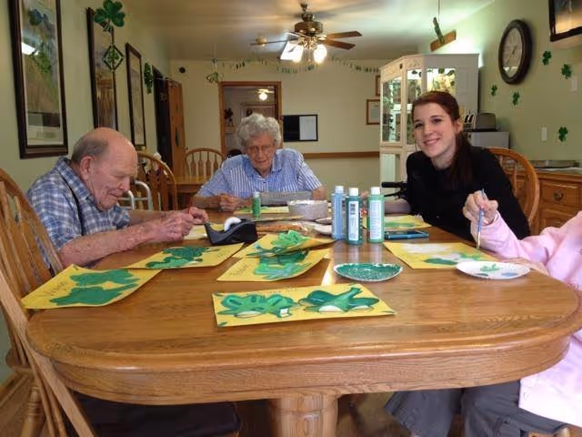 Four people seated around a wooden table painting green shamrock crafts in a decorated communal dining room.