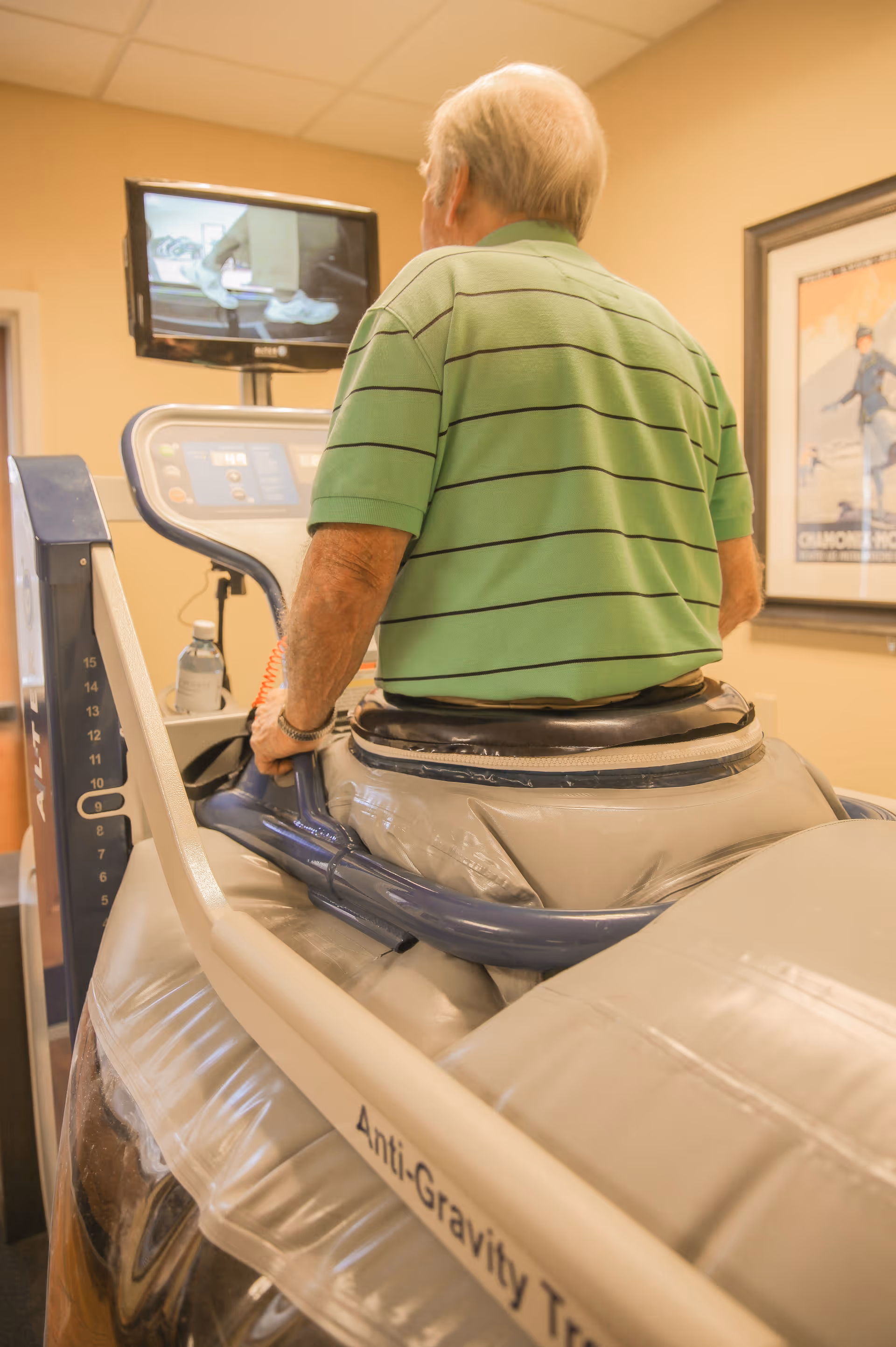 An elderly man wearing a green striped polo shirt is using an anti-gravity treadmill in a fitness or rehabilitation room. A monitor in front of him displays a close-up of his feet walking on the treadmill. The room has beige walls and a framed picture on the wall.