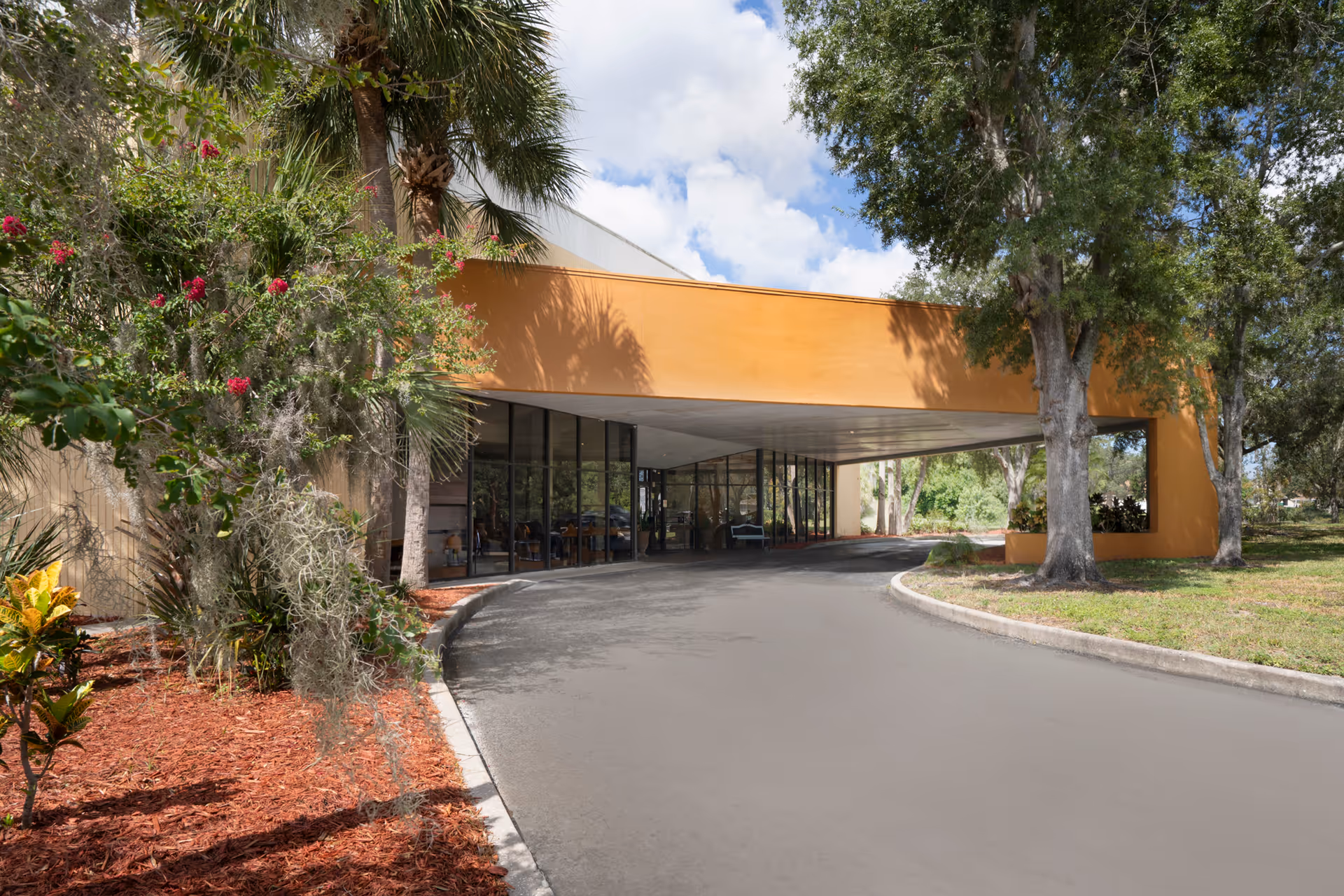 Exterior view of the entrance to Peninsula Health Center featuring a covered driveway with large glass windows, surrounded by trees and landscaped greenery under a partly cloudy sky.