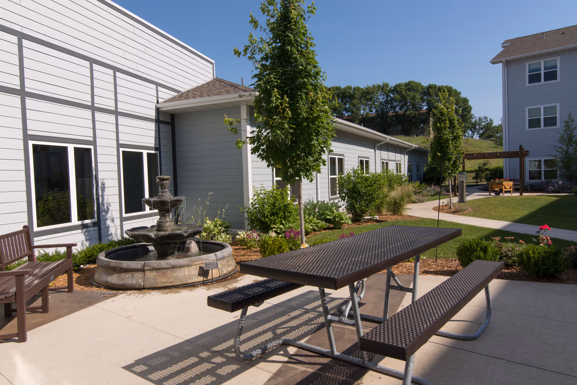 Outdoor courtyard at a retirement community with a picnic table, fountain, benches, landscaping, and adjacent buildings.