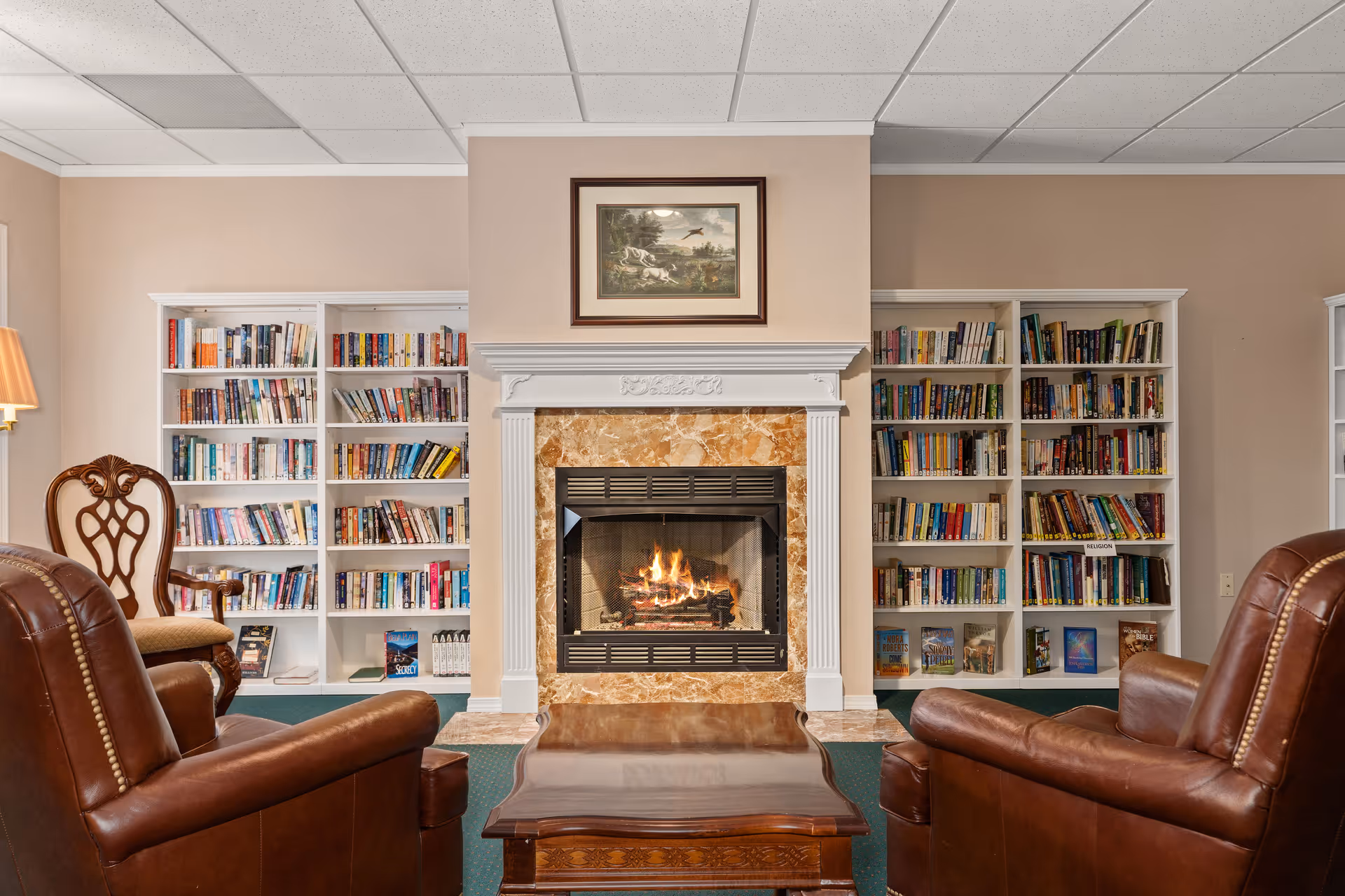 Cozy common room with a lit fireplace flanked by bookshelves and leather chairs facing a coffee table.