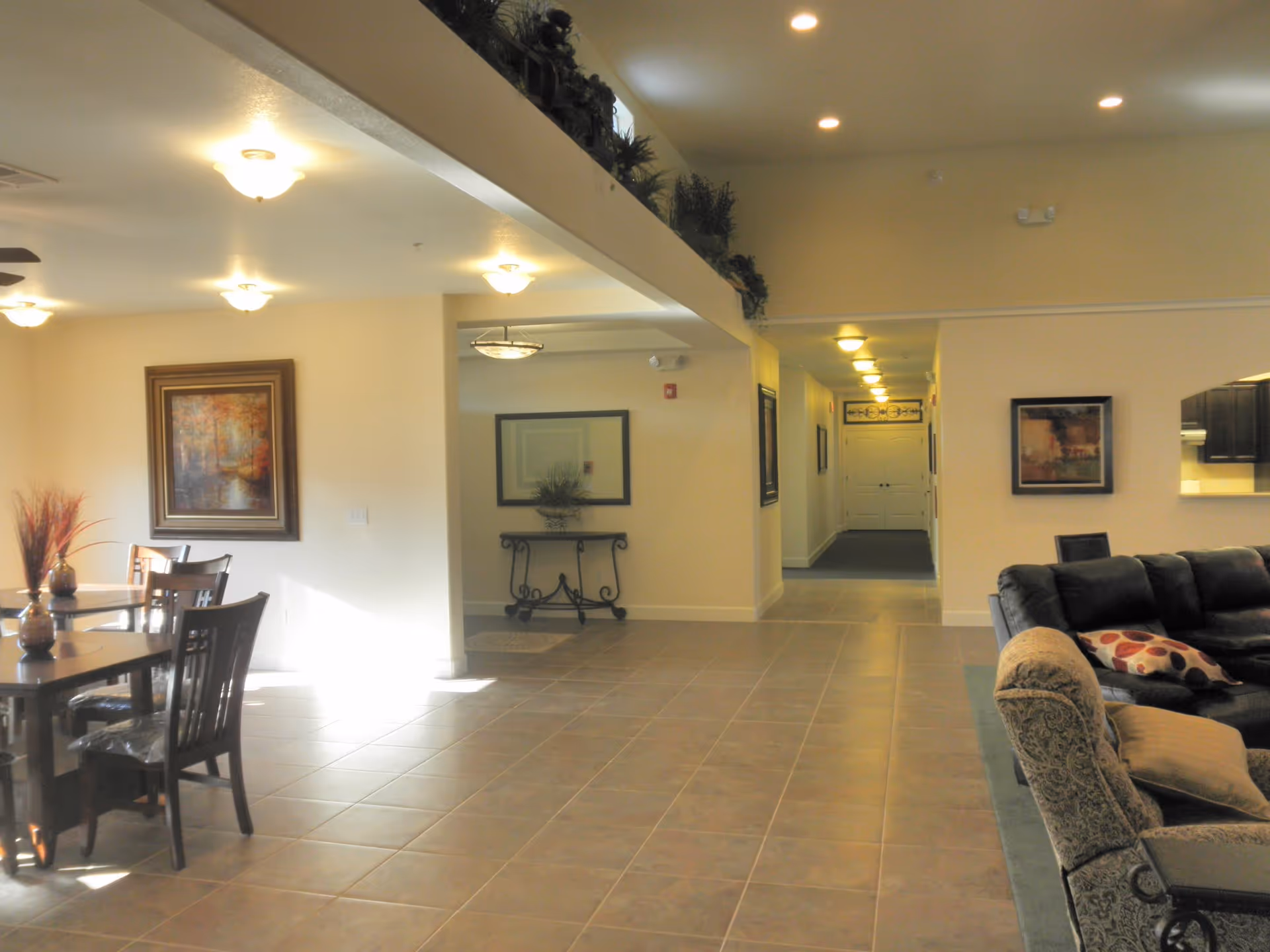 Interior view of a senior living facility common area with tiled floor, a dining table with chairs on the left, a hallway in the center, and a living room area with a sofa and armchair on the right. The walls are decorated with framed artwork and there are ceiling lights providing illumination.