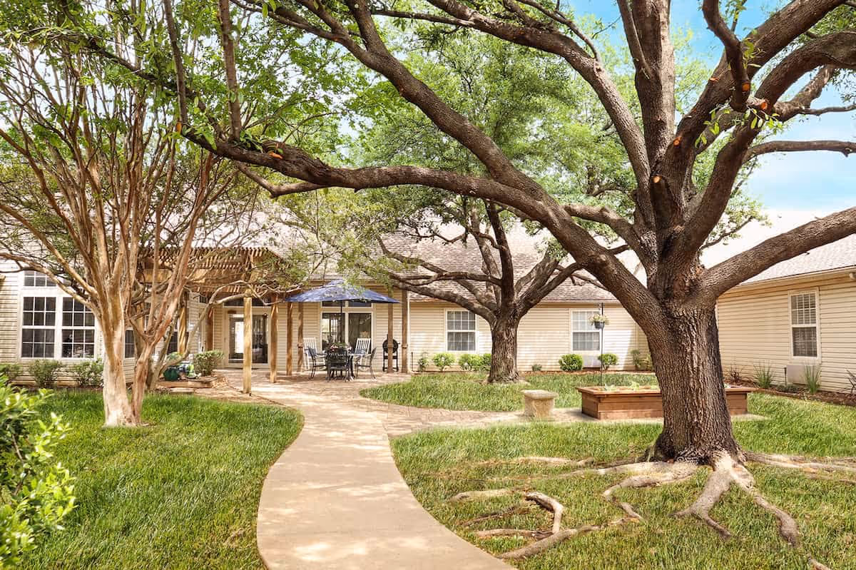 A landscaped courtyard with a winding paved path, large shade trees, patio seating under an umbrella, and surrounding single-story building.