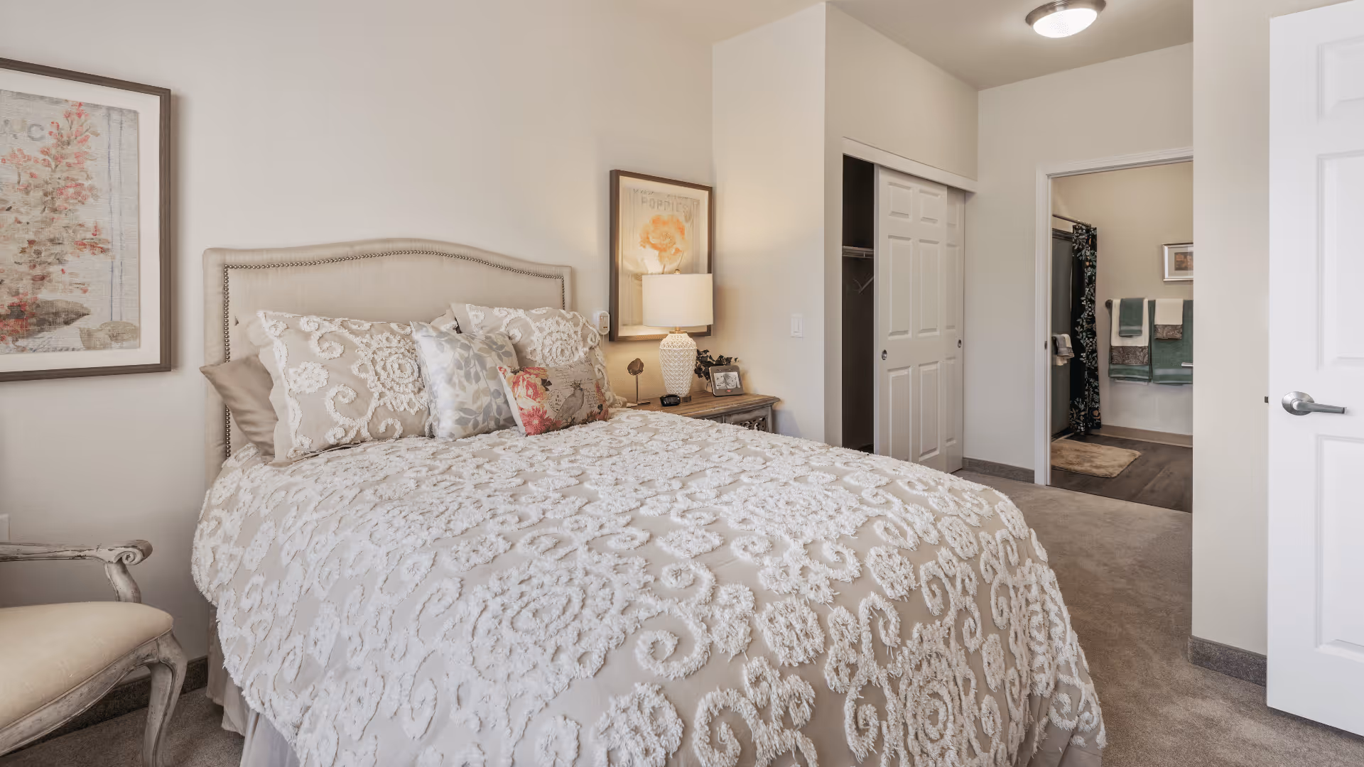 A cozy bedroom with a beige upholstered bed covered in a textured white and beige bedspread, multiple decorative pillows, a wooden nightstand with a lamp and framed artwork on the wall. There is a closet with sliding doors and an open doorway leading to a bathroom with towels hanging on the wall.