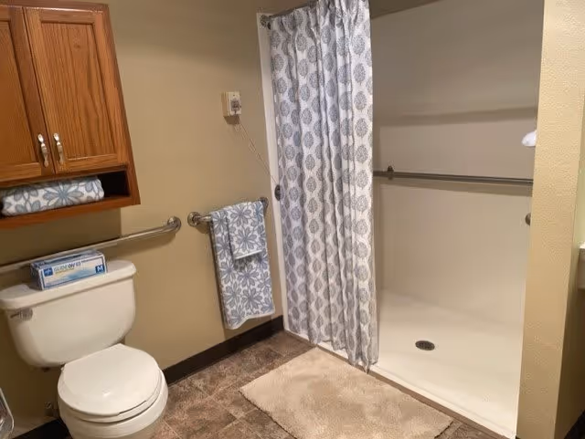 A bathroom with a white toilet, a wooden cabinet above it, a towel rack with a patterned blue and white towel, and a walk-in shower with a patterned shower curtain. The floor has brown tiles and there is a beige bath mat in front of the shower.