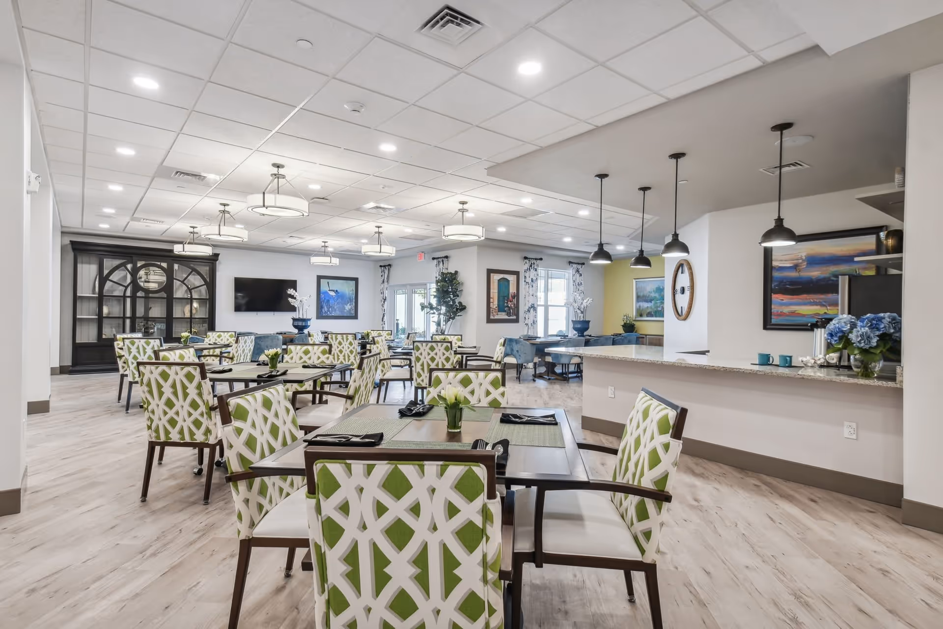 A spacious and well-lit dining area in a senior living facility with multiple tables and chairs. The chairs have a green and white geometric pattern. The room features modern ceiling lights, a large flat-screen TV mounted on the wall, decorative paintings, and a counter with pendant lights hanging above it. There are also some plants and floral arrangements adding to the decor.