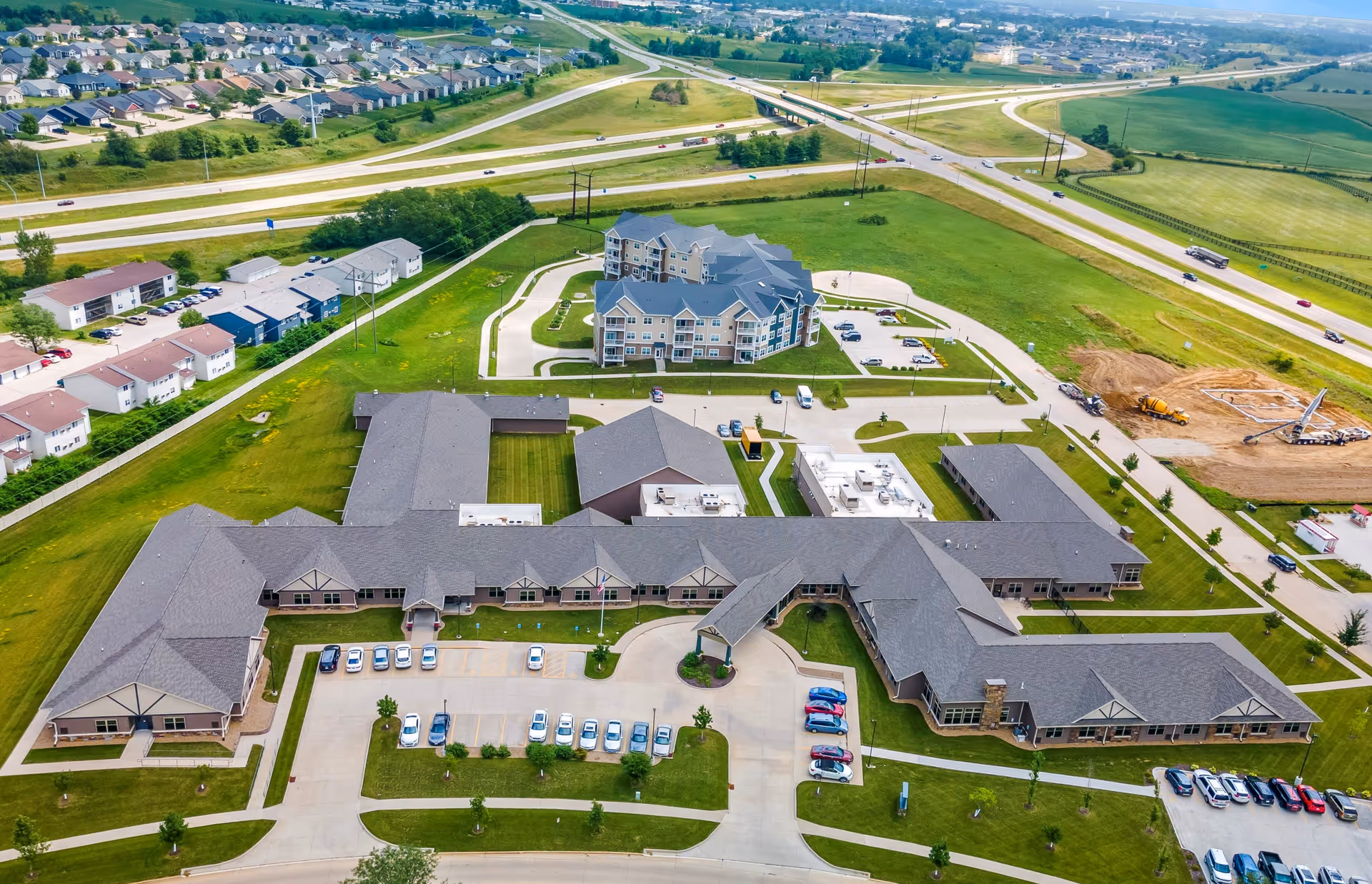 Aerial view of The Gardens of Cedar Rapids senior living facility showing multiple buildings with parking lots, green lawns, and surrounding roads and highways in a suburban area.