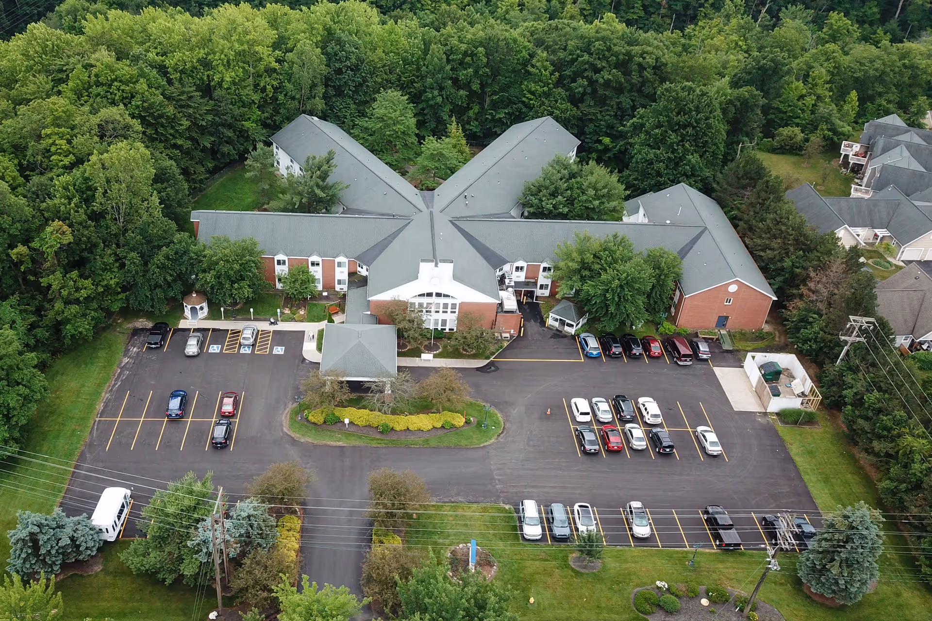 Aerial view of a senior living facility surrounded by trees and greenery. The building has a unique star-shaped layout with multiple wings and a large parking lot with several cars parked. The facility is set in a suburban area with neighboring houses visible on one side.