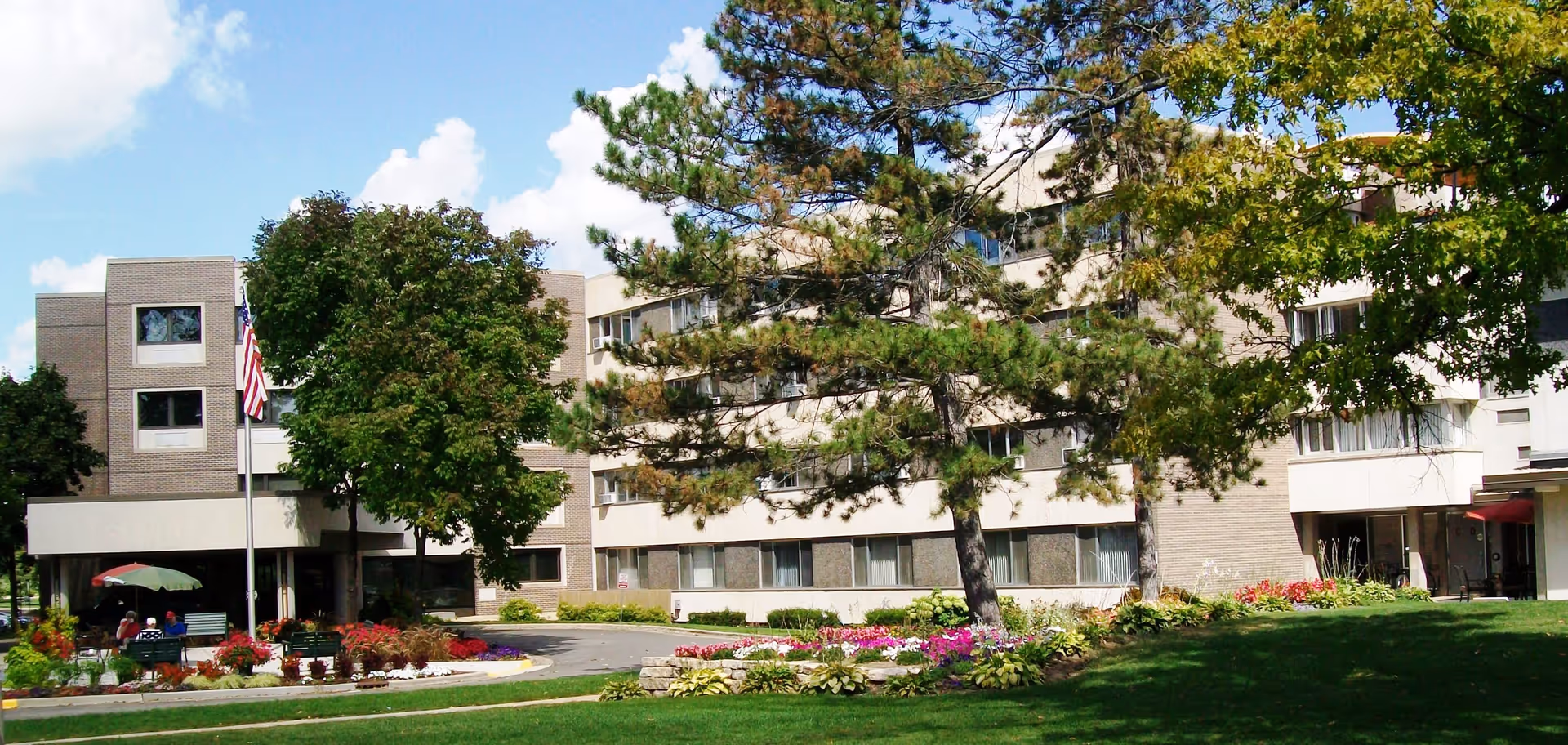 Exterior view of a multi-story senior living facility building surrounded by green trees, colorful flower beds, and a well-maintained lawn under a partly cloudy blue sky. There is an American flag on a flagpole near the entrance and a few people sitting on benches under an umbrella.