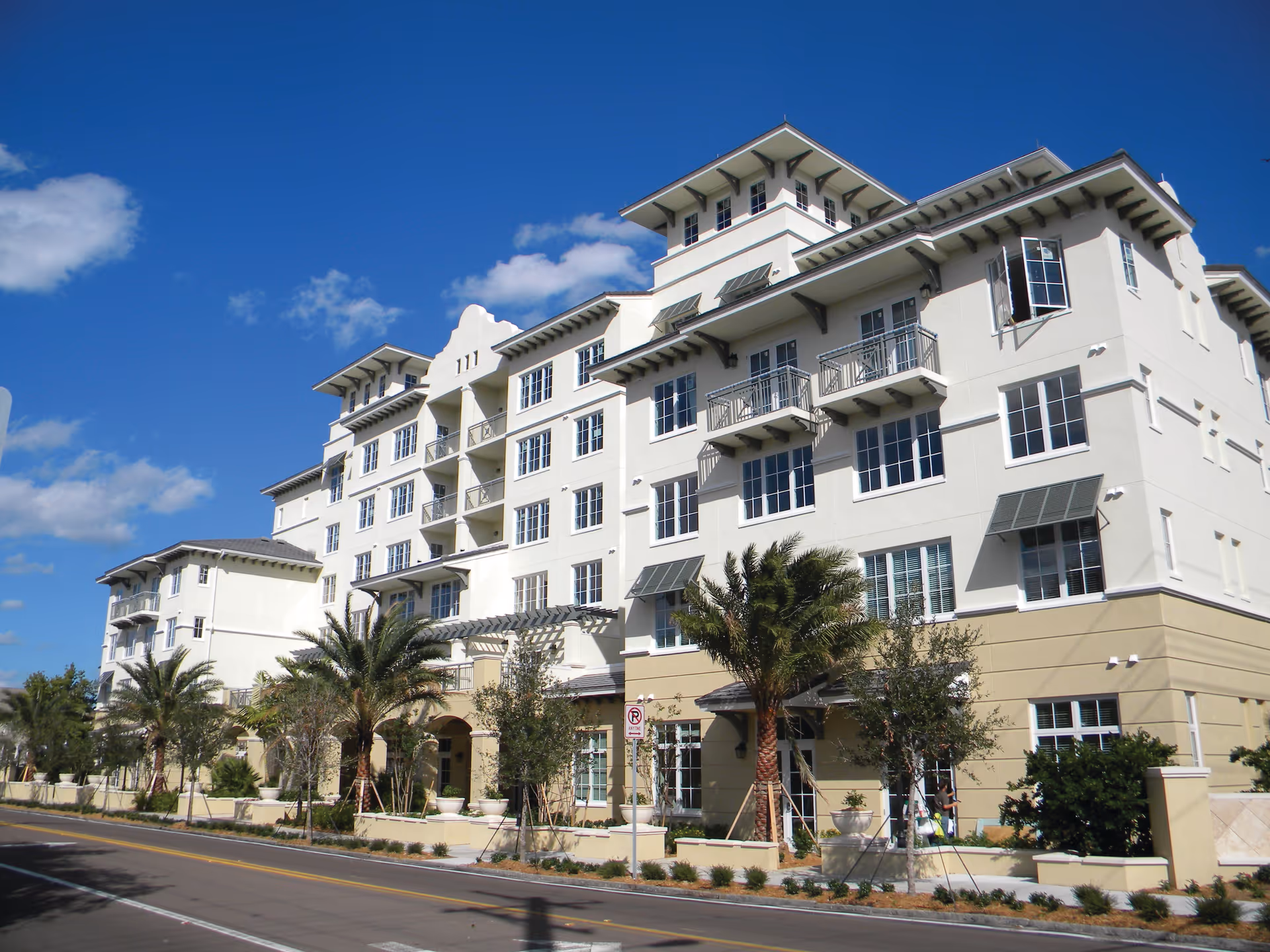 Front exterior of a multi-story white senior living building with balconies, palm trees, and a clear blue sky.