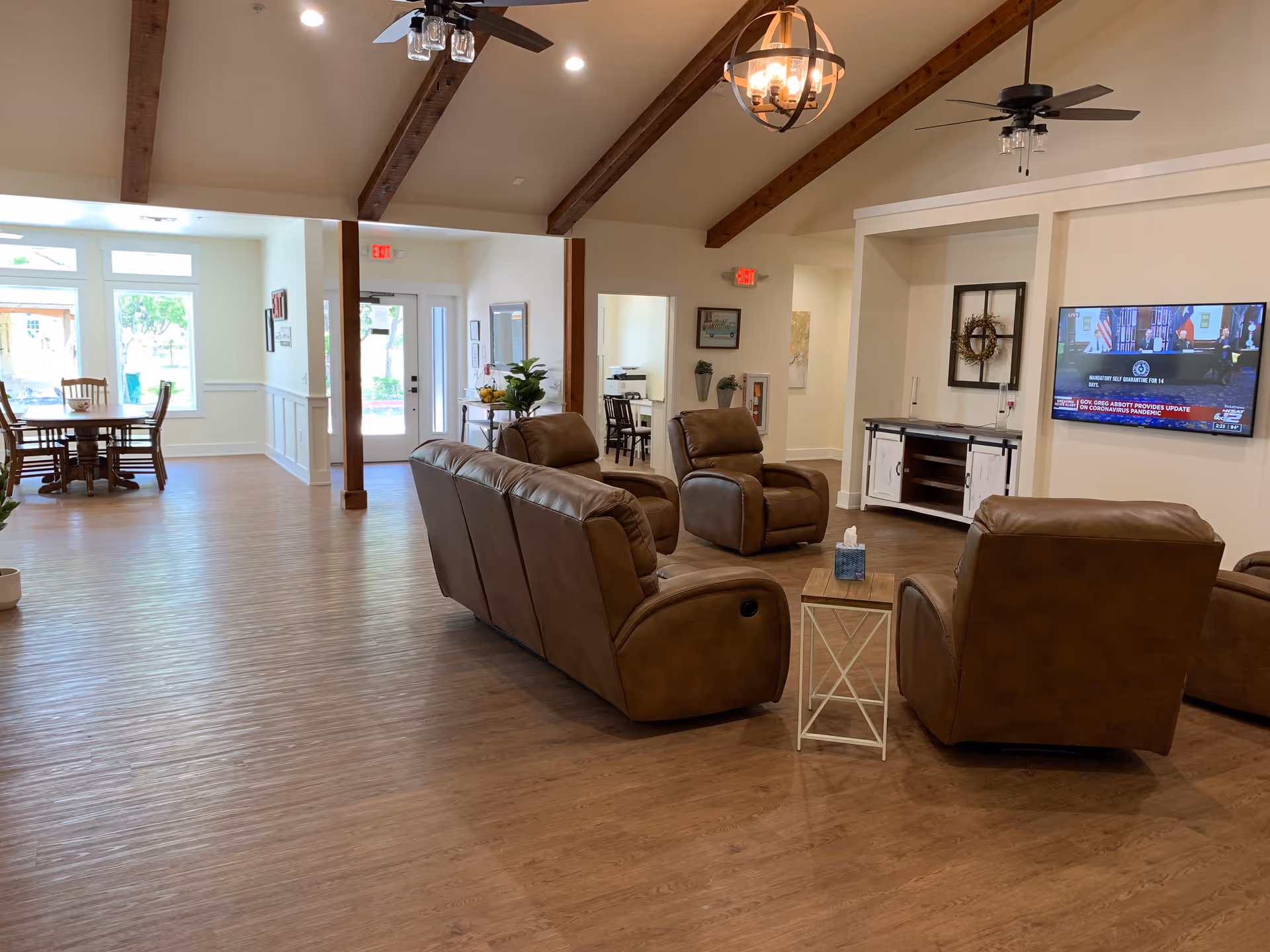 A spacious living room area in an assisted living facility with brown leather recliners arranged around a small wooden side table. A flat-screen TV is mounted on the wall showing a news broadcast. The room features wooden beams on a vaulted ceiling, ceiling fans with lights, and wood flooring. In the background, there is a dining area with a wooden table and chairs near large windows letting in natural light.
