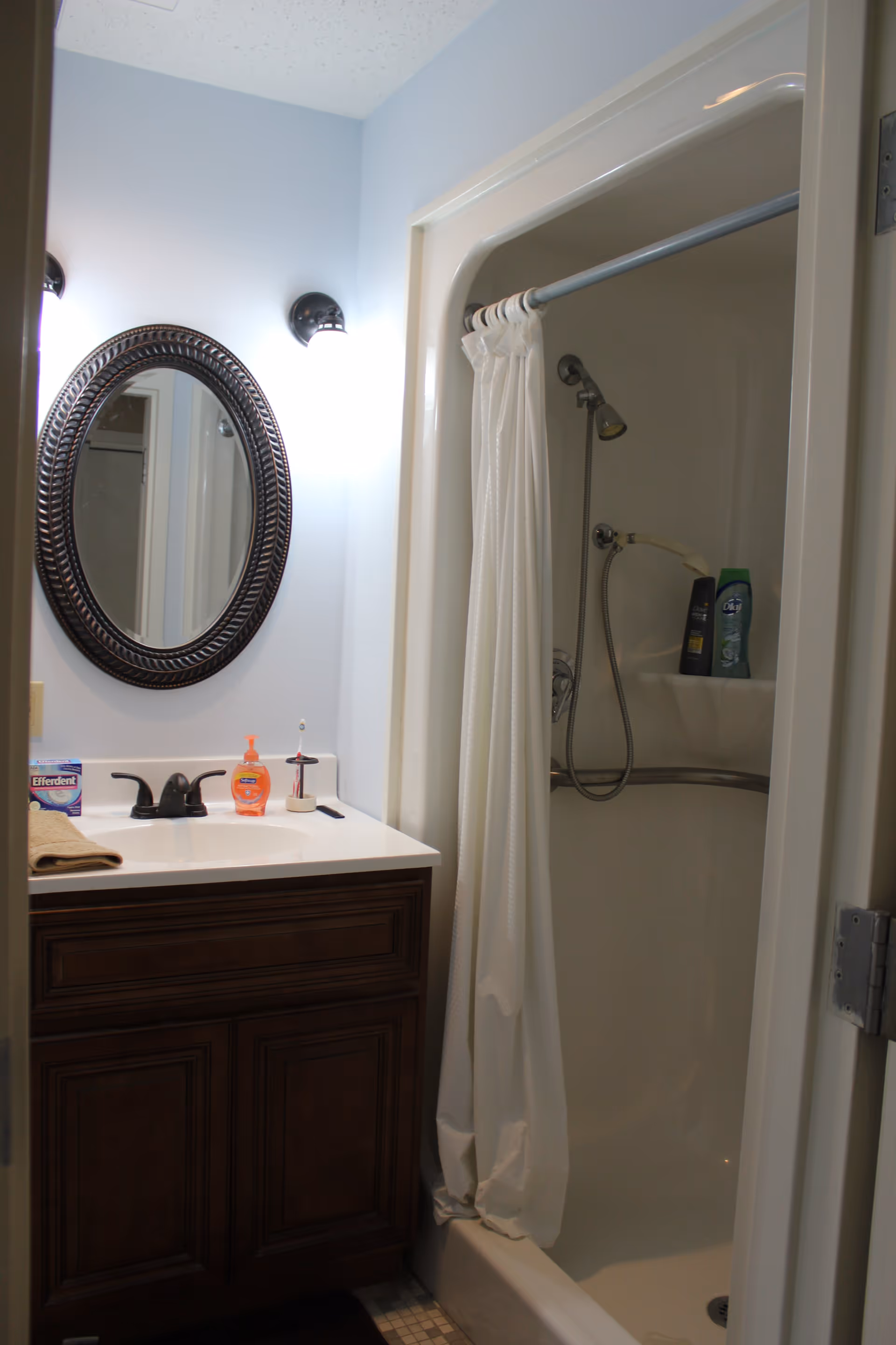 A small bathroom with a dark wooden vanity topped with a white sink and black faucet. Above the sink is an oval mirror with a decorative frame and two wall-mounted lights. To the right is a shower with a white curtain partially drawn, a handheld showerhead, and two bottles of shower products on a built-in shelf.
