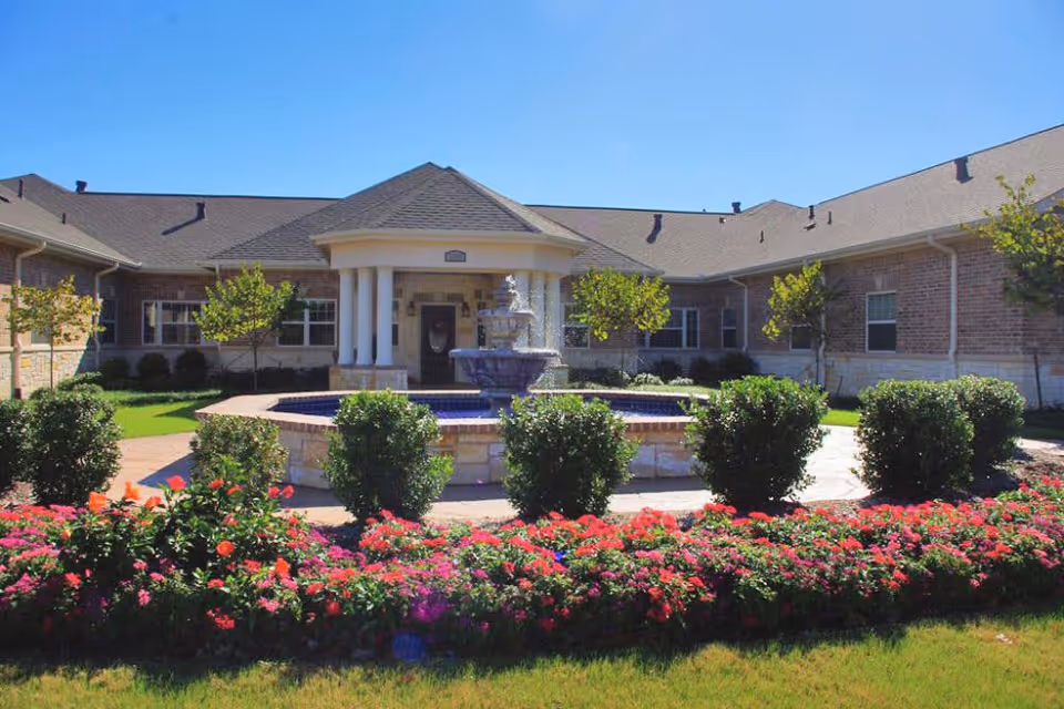 Exterior view of Oak Ridge Alzheimer's Special Care Center showing a single-story brick building with a central entrance featuring white columns and a peaked roof. In front of the entrance is a circular stone fountain surrounded by neatly trimmed bushes and vibrant flower beds under a clear blue sky.