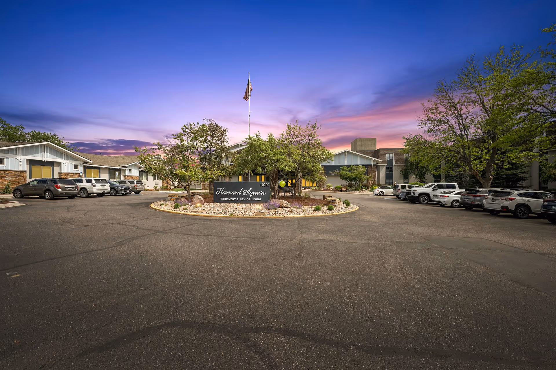Exterior view of Harvard Square By Cogir senior living facility at dusk, showing a circular driveway with parked cars, landscaped center island with trees and a sign reading 'Harvard Square Retirement & Senior Living', and buildings in the background under a colorful sky.