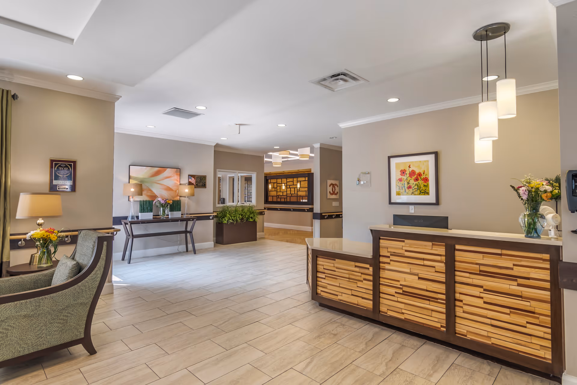 A bright and spacious reception area in Stratford Commons Rehabilitation & Health Care featuring a wooden front desk with pendant lights above, a framed floral painting on the wall, comfortable seating with green upholstered chairs, side tables with lamps and flower vases, a console table with lamps and plants, and decorative wall art in a neutral color scheme.