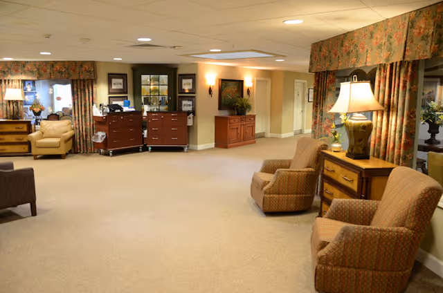 A spacious and well-lit common area in a senior living facility with beige carpet, comfortable armchairs, wooden side tables with lamps, floral curtains, and decorative wall art. There are cabinets and a reception desk area in the background.