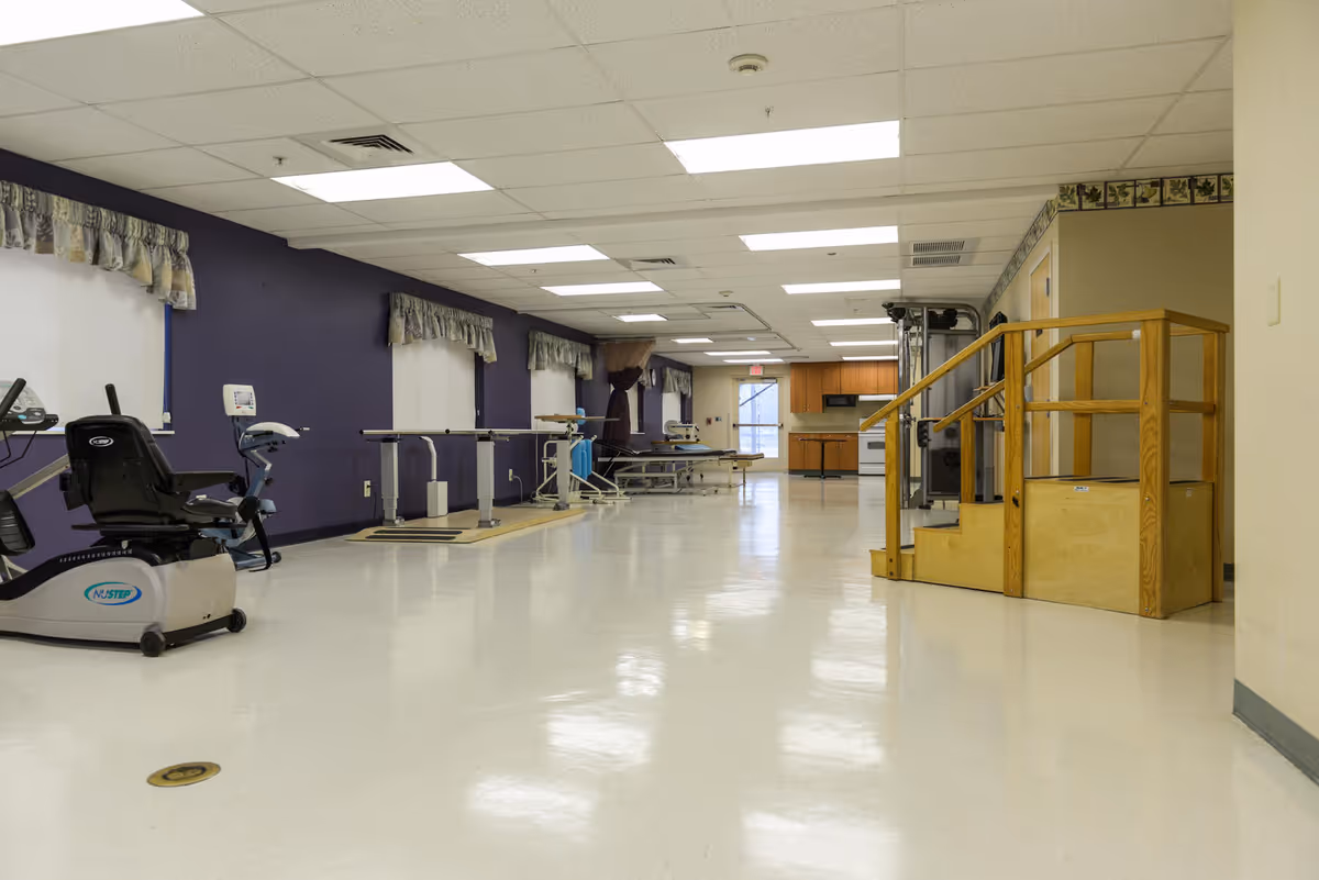 A spacious rehabilitation room with exercise equipment including a recumbent bike, parallel bars for walking practice, therapy tables, and a wooden staircase with handrails for physical therapy. The room has a white floor, purple walls with windows covered by valances, and a kitchenette area in the background.