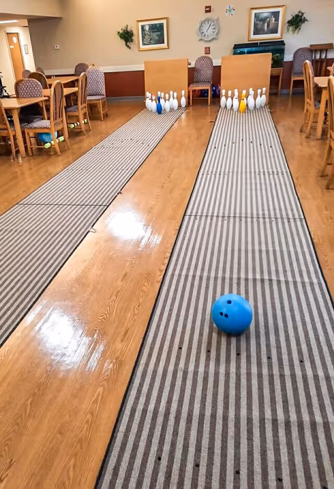 Indoor senior activity/dining room with two makeshift bowling lanes, a blue bowling ball in the foreground and pins set at the far end between tables and chairs.