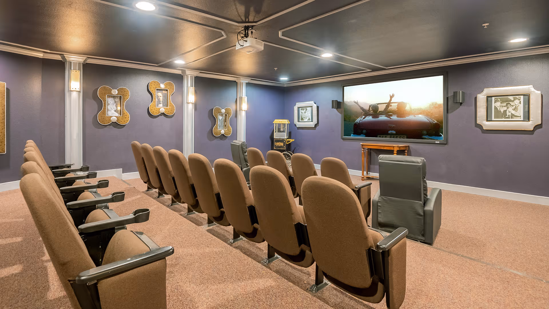 A small theater room with two rows of brown cushioned seats and two gray recliners facing a large screen showing a car with two people raising their hands. The walls are painted purple and decorated with framed pictures and wall sconces. There is a popcorn machine in the corner.