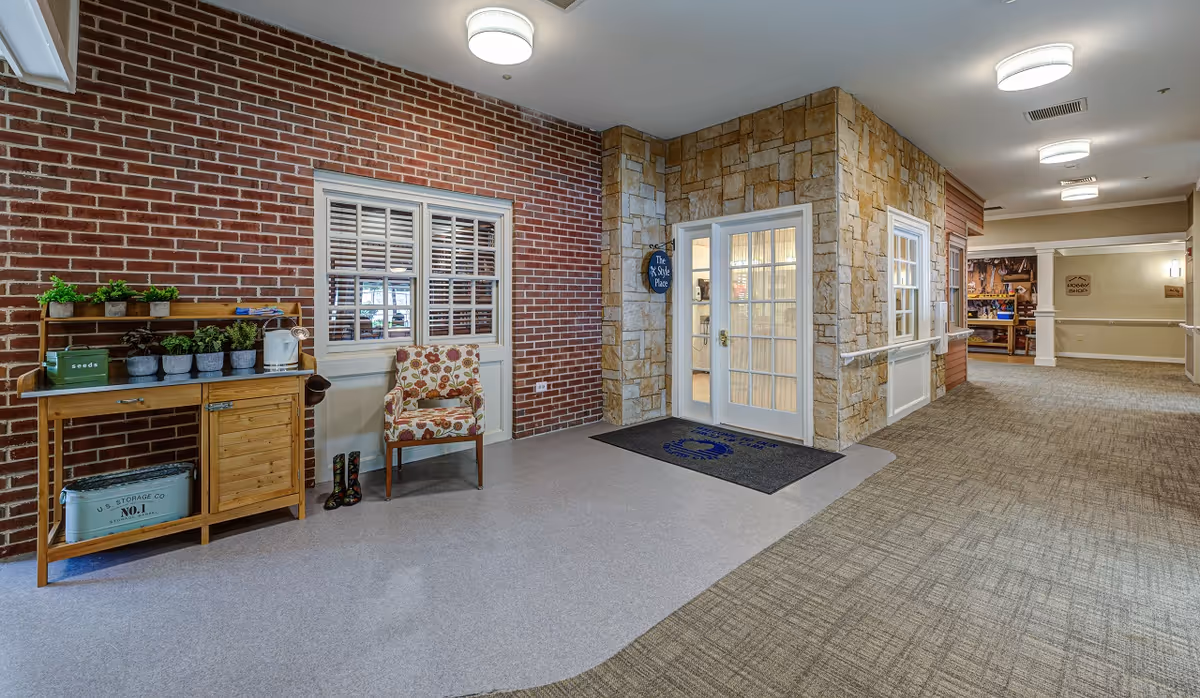 Interior hallway of a senior living facility with a brick wall on the left featuring a wooden table with small potted plants and gardening items, a floral upholstered chair, and a pair of rain boots. To the right, there is a stone wall with a glass door entrance labeled 'The Style Place' and a carpeted hallway extending into the distance with ceiling lights.