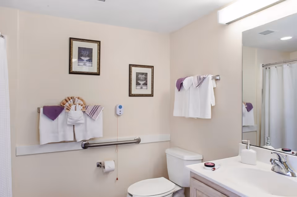 A clean and well-lit bathroom featuring a toilet, a sink with a faucet, a large mirror, and a shower with a white curtain. The walls are beige, decorated with two framed pictures. There are white and purple towels neatly hung on towel racks, and a grab bar is installed next to the toilet for support.