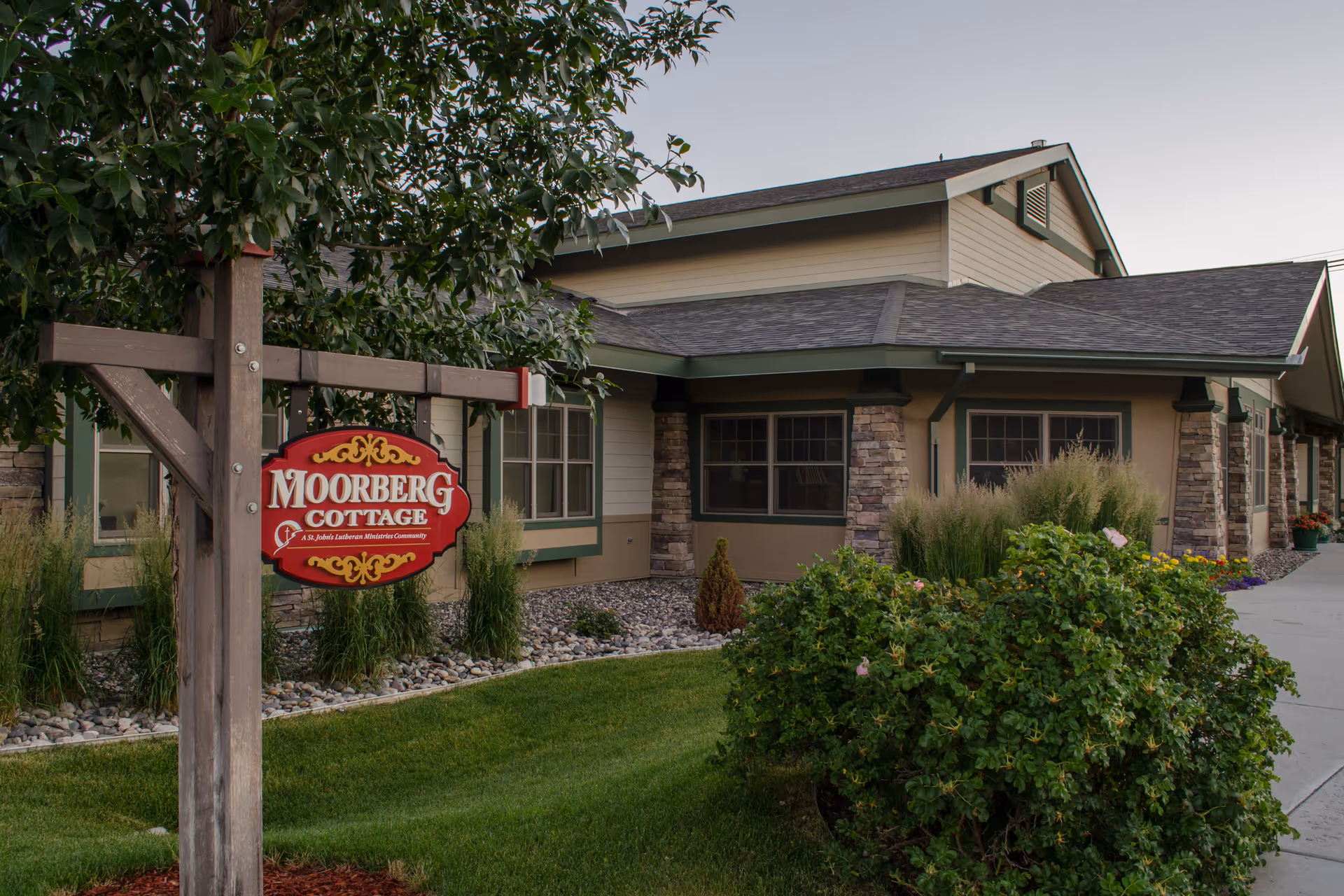 Exterior view of a single-story building with beige siding, stone accents, and green trim. In front of the building is a wooden post with a red sign that reads 'Moorberg Cottage, A St. John's Lutheran Ministries Community'. There are bushes, grass, and a concrete walkway visible around the building.