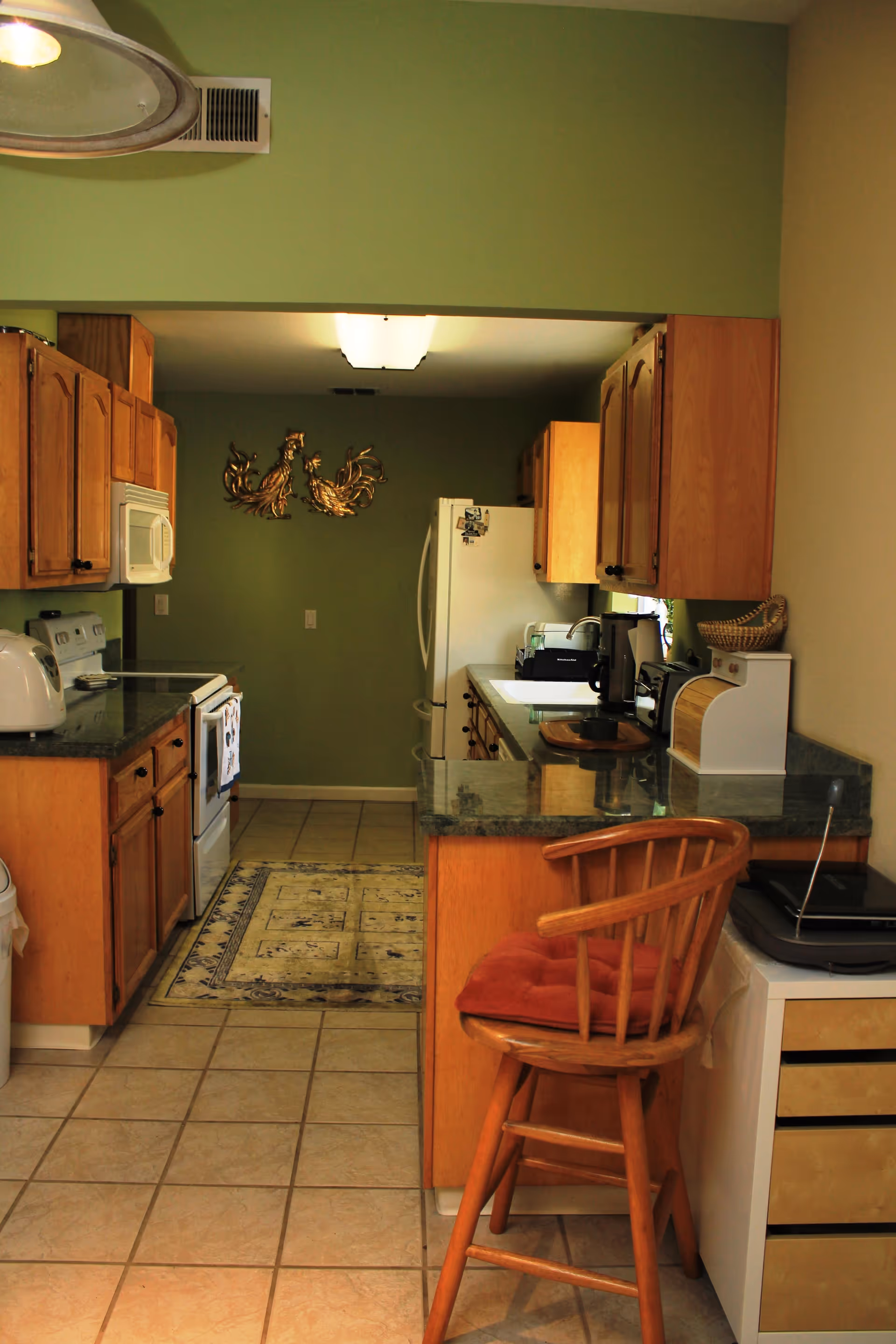 Galley-style kitchen with wooden cabinets, green walls and countertops, white appliances, and a wooden bar stool at a breakfast counter.