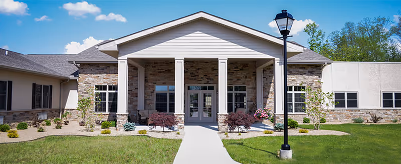 Front exterior view of a single-story building with a covered entrance supported by four white columns, stone and beige siding, a concrete walkway leading to double glass doors, landscaped bushes and small trees, and a black street lamp on the right side of the walkway.