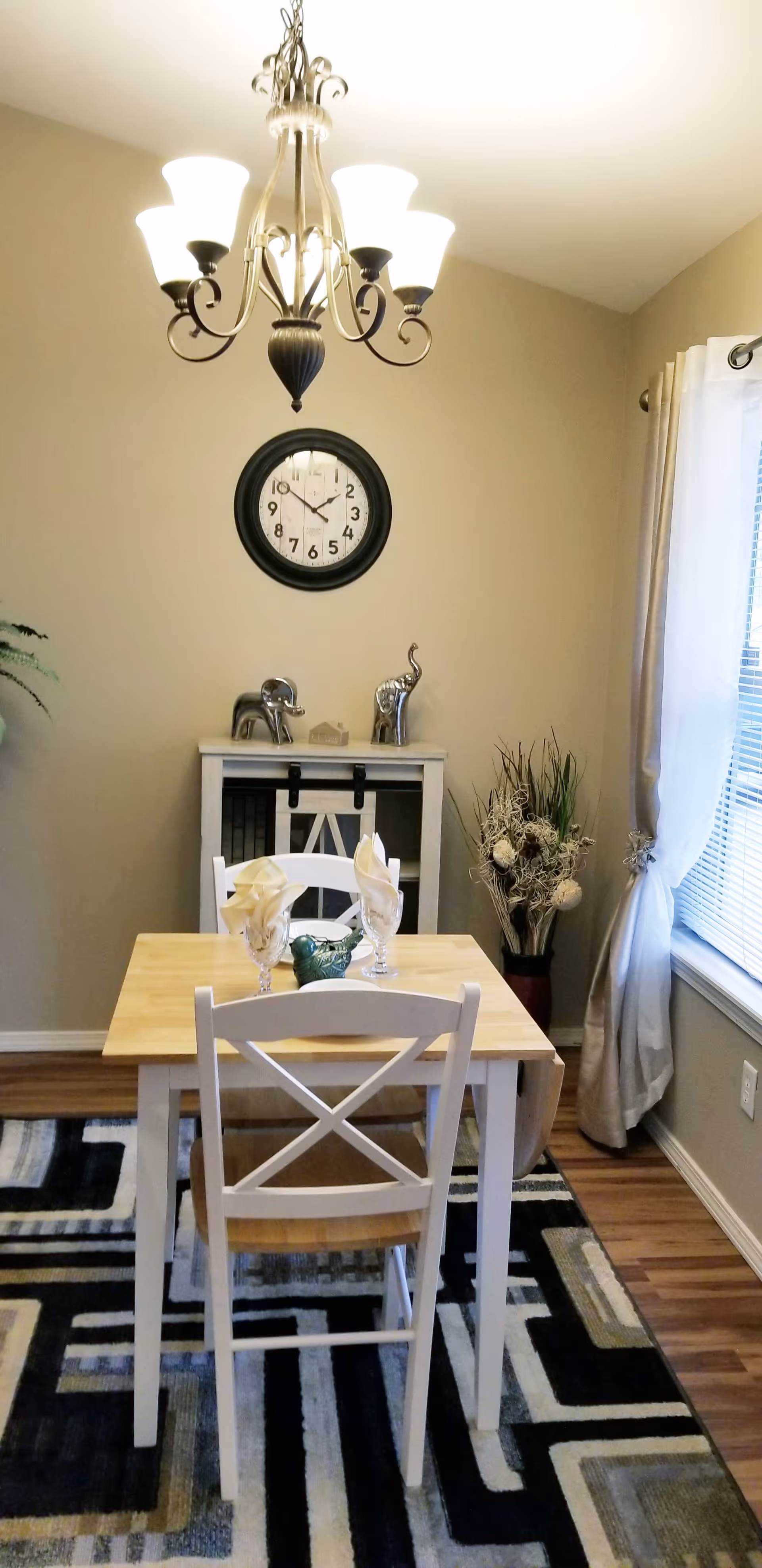 A small dining area with a wooden table set for two, featuring two white chairs with cross-back design. Above the table hangs a decorative chandelier with five lights. On the wall behind the table is a round black clock showing the time as 11:10. Below the clock is a small white cabinet with two silver elephant figurines on top. To the right of the cabinet is a tall vase with dried decorative plants. A window with white curtains is on the right side of the room, and the floor has a patterned area rug with black, white, and gray geometric designs.