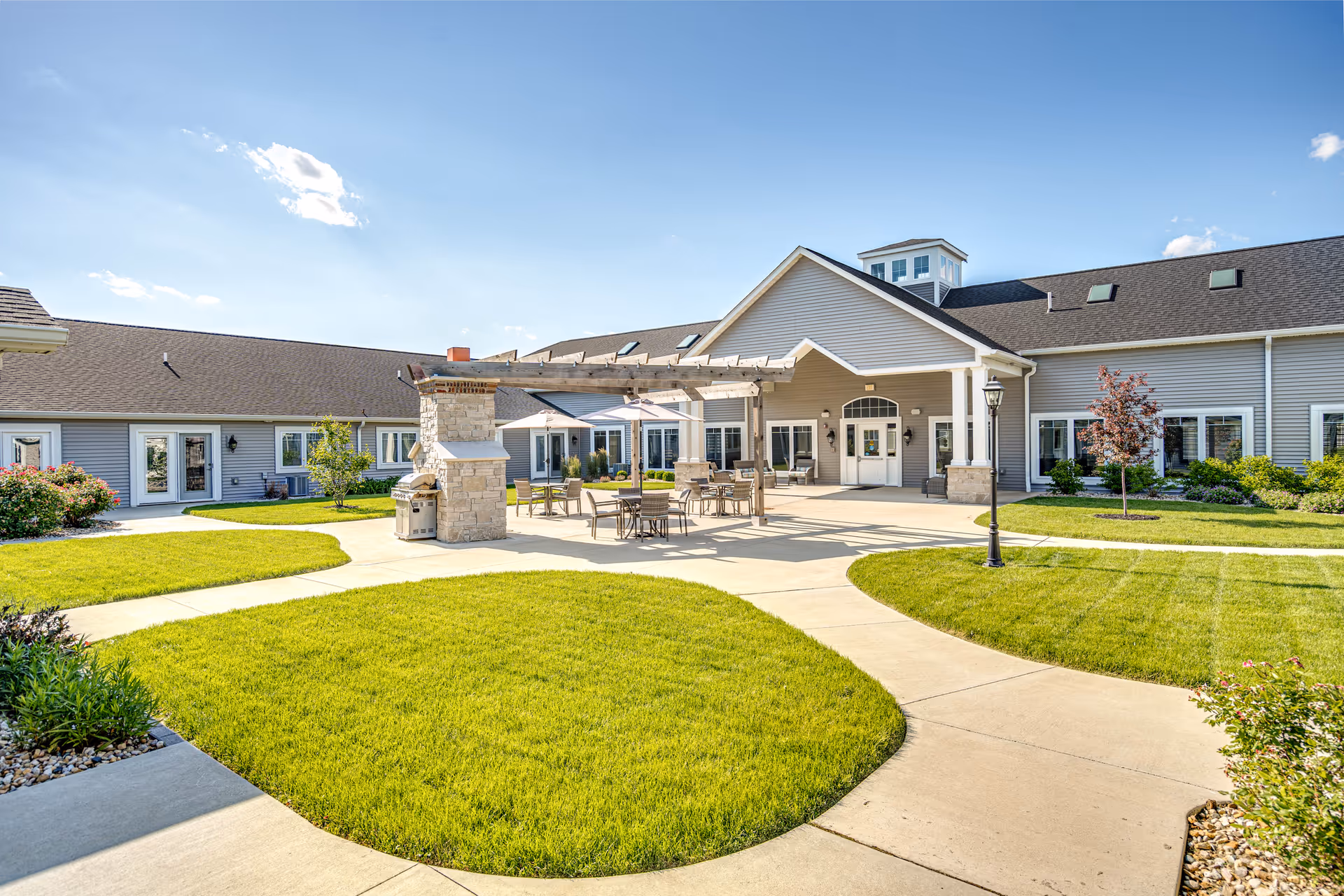 Outdoor courtyard area of Villas of Holly Brook Assisted Living in Washington, IL, featuring a paved walkway, green lawns, a pergola with tables and chairs underneath, and the building exterior with multiple windows and doors under a clear blue sky.