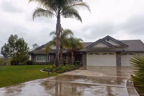 Wet driveway and palm trees frame a single-story home with a two-car garage.