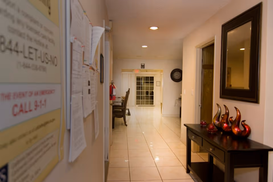 A well-lit hallway with tiled floors and beige walls. On the left wall, there are various papers and notices pinned up. On the right side, there is a dark wooden console table with decorative red and orange glass sculptures and a large framed mirror above it. At the end of the hallway, there are chairs and a glass door with vertical blinds. A clock is mounted on the far wall.